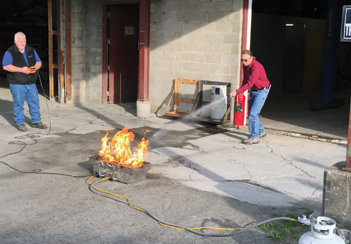John Hollinrake, an ARC estimator, puts out the flashpan fire at the fire extinguisher station. The flashpan is connected to a propane tank and sensor, which the instructor, Carle Underhill, Catamount Safety, is holding.